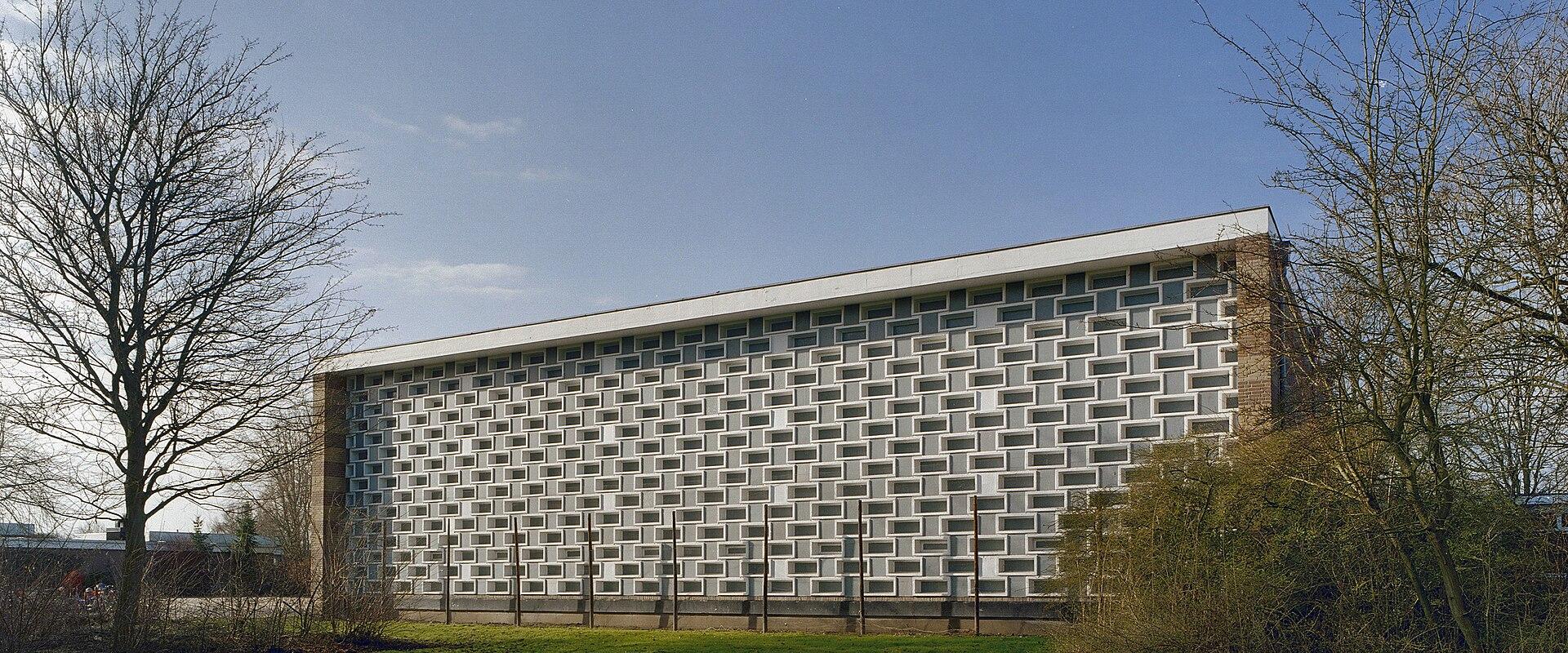 A long, low-rise modernist building with a distinctive white facade featuring a grid of recessed rectangular windows, set against a blue sky with bare trees and green grass in the foreground.