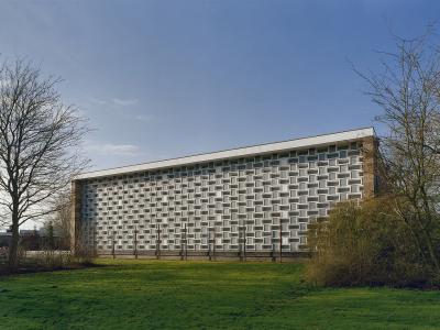 A long, low-rise modernist building with a distinctive white facade featuring a grid of recessed rectangular windows, set against a blue sky with bare trees and green grass in the foreground.