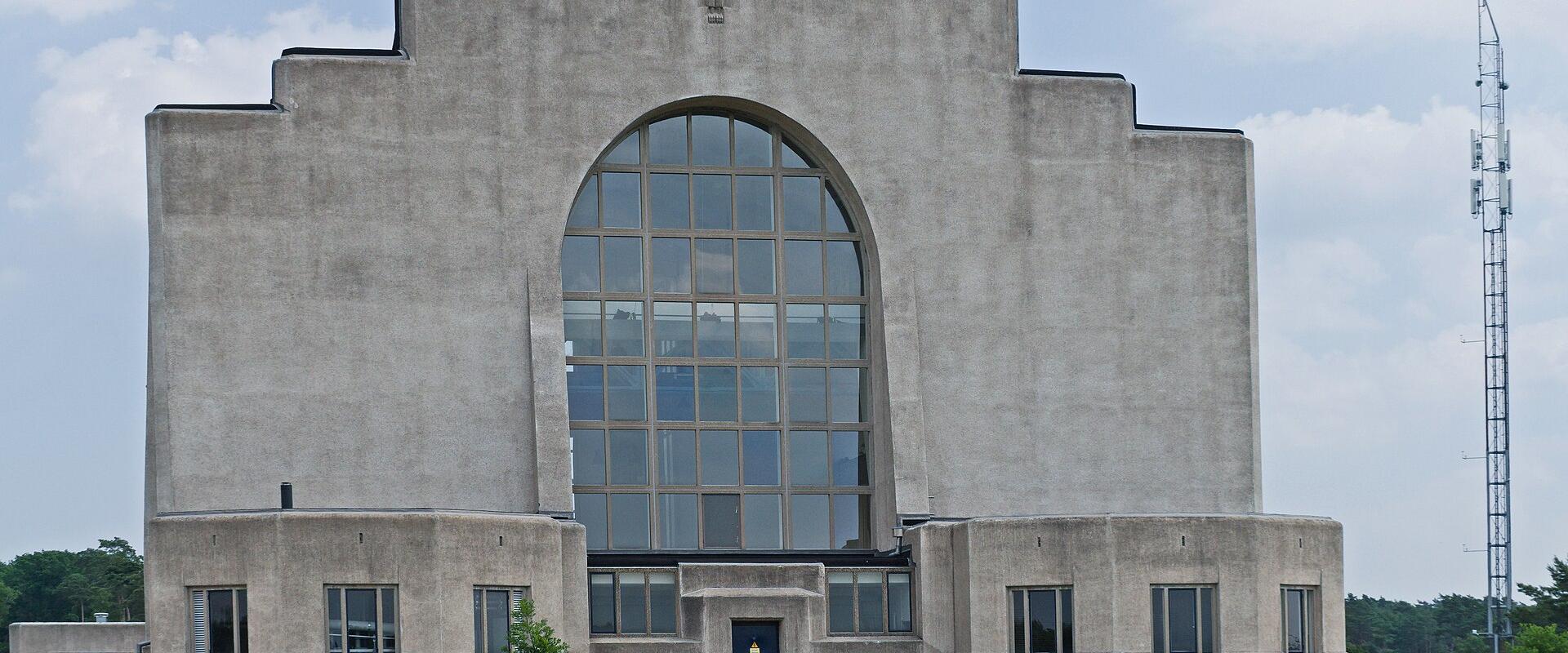 A large, grey Art Deco concrete building with a prominent central arched window and stepped roofline, situated in a grassy heathland setting with a tall radio mast visible to the right.
