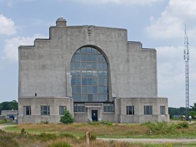A large, grey Art Deco concrete building with a prominent central arched window and stepped roofline, situated in a grassy heathland setting with a tall radio mast visible to the right.