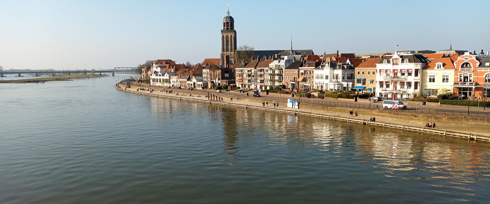 A wide-angle view of the riverfront in Deventer, Netherlands, featuring the distinctive tower of the Grote Kerk (St. Lebuïnus Church) and a row of historic buildings along the IJssel riverbank.