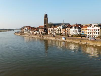 A wide-angle view of the riverfront in Deventer, Netherlands, featuring the distinctive tower of the Grote Kerk (St. Lebuïnus Church) and a row of historic buildings along the IJssel riverbank.