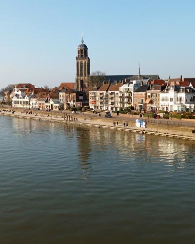A wide-angle view of the riverfront in Deventer, Netherlands, featuring the distinctive tower of the Grote Kerk (St. Lebuïnus Church) and a row of historic buildings along the IJssel riverbank.