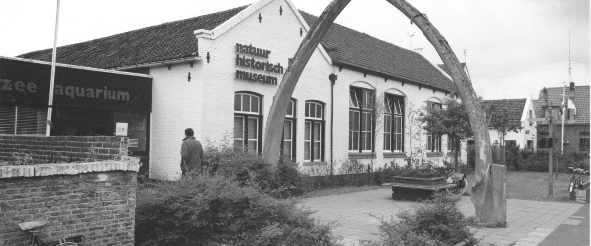 A black-and-white photograph showing a white brick building with the text "natuur historisch museum" on its facade, flanked by a large whale jawbone arch and an adjacent "zee aquarium" sign.