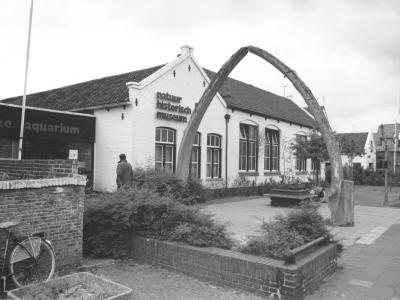 A black-and-white photograph showing a white brick building with the text "natuur historisch museum" on its facade, flanked by a large whale jawbone arch and an adjacent "zee aquarium" sign.