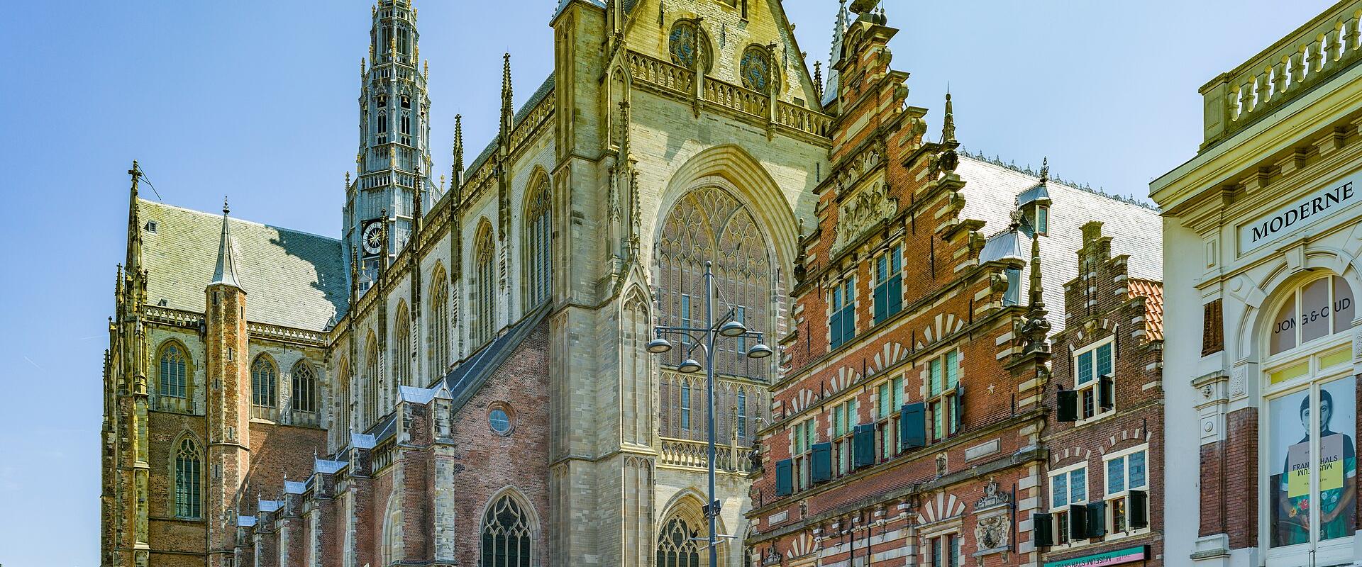 The image shows the St. Bavokerk (Grote Kerk) in Haarlem, Netherlands, featuring its distinctive late Gothic architecture, tall spire, and adjacent historic buildings on the Grote Markt square.