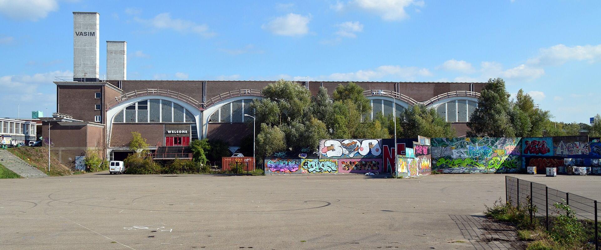 A large, brown brick industrial building with two tall concrete chimneys labeled "VASIM" and a series of arched windows, fronted by a paved lot and a wall covered in graffiti.