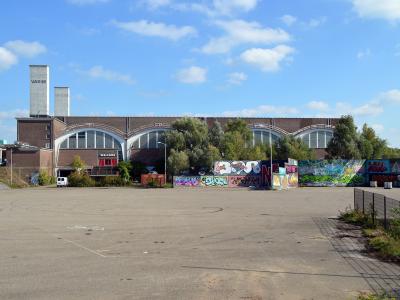 A large, brown brick industrial building with two tall concrete chimneys labeled "VASIM" and a series of arched windows, fronted by a paved lot and a wall covered in graffiti.