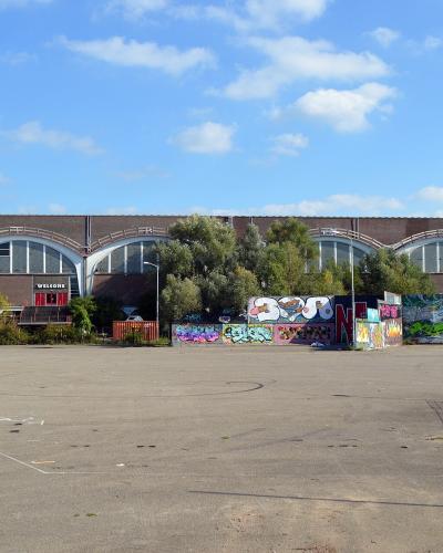 A large, brown brick industrial building with two tall concrete chimneys labeled "VASIM" and a series of arched windows, fronted by a paved lot and a wall covered in graffiti.