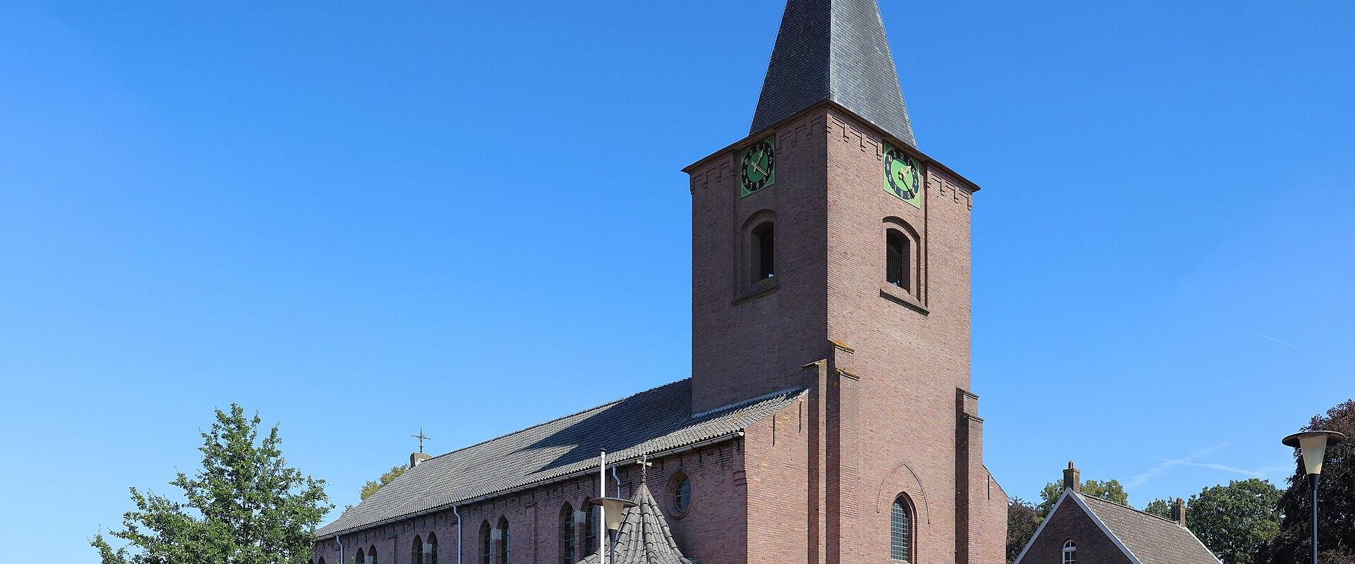 A red brick church with a tall, pointed steeple and green clock faces stands on a paved town square under a clear blue sky.