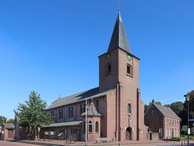 A red brick church with a tall, pointed steeple and green clock faces stands on a paved town square under a clear blue sky.