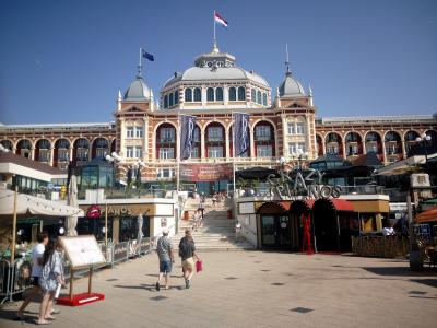 A large, ornate red-brick and white stone building with a central dome and arched windows, featuring vertical banners with the text "KURHAUS" and a large "CRAZY PIANOS" sign in the foreground plaza.