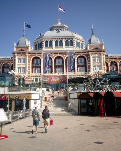 A large, ornate red-brick and white stone building with a central dome and arched windows, featuring vertical banners with the text "KURHAUS" and a large "CRAZY PIANOS" sign in the foreground plaza.