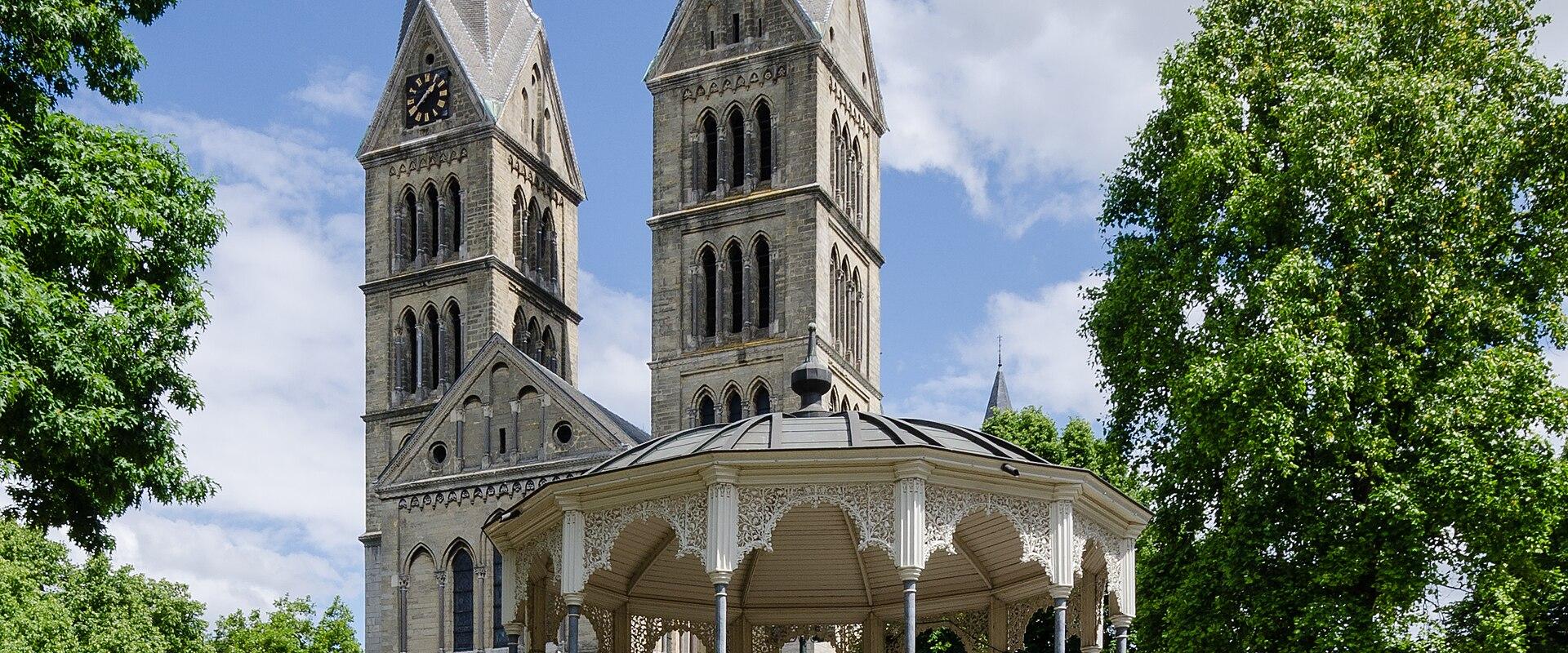 A large, historic Romanesque-style cathedral with multiple towers and spires, situated in an urban setting with a street and parked car in the foreground.