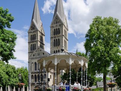 A large, historic Romanesque-style cathedral with multiple towers and spires, situated in an urban setting with a street and parked car in the foreground.