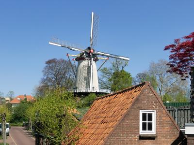 A white smock windmill with a brick base stands on a hill behind a brick building with a red-tiled roof, under a clear blue sky.