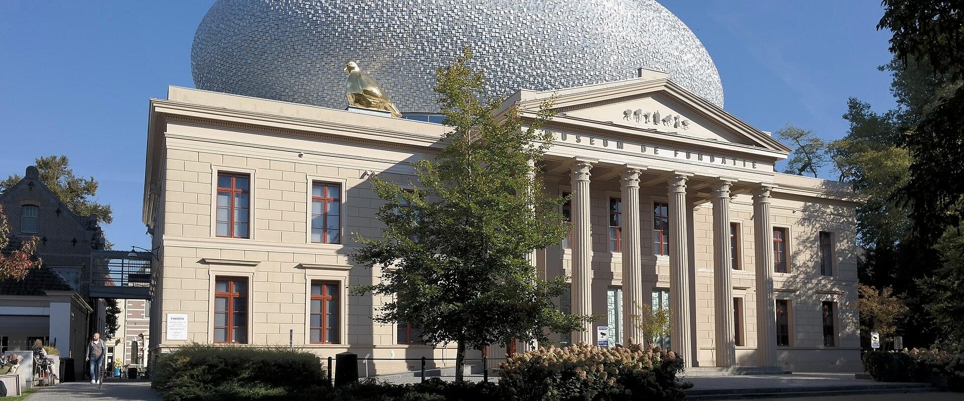 A neoclassical building with a large silver dome and a golden sculpture on the roof, with "MUSEUM DE FUNDATIE" inscribed on the pediment.