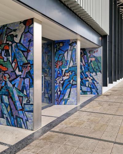 A view of the exterior colonnade of a modern building featuring large, blue-toned abstract mosaic murals on the pillars, with a paved walkway and a distinctive metal lattice roof structure overhead.