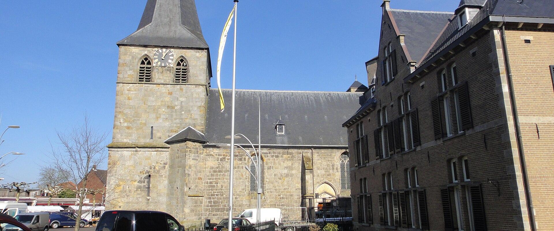 A stone church with a tall, pointed spire and clock tower stands next to a brick building under a blue sky, with cars parked in the foreground.