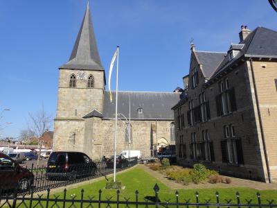 A stone church with a tall, pointed spire and clock tower stands next to a brick building under a blue sky, with cars parked in the foreground.