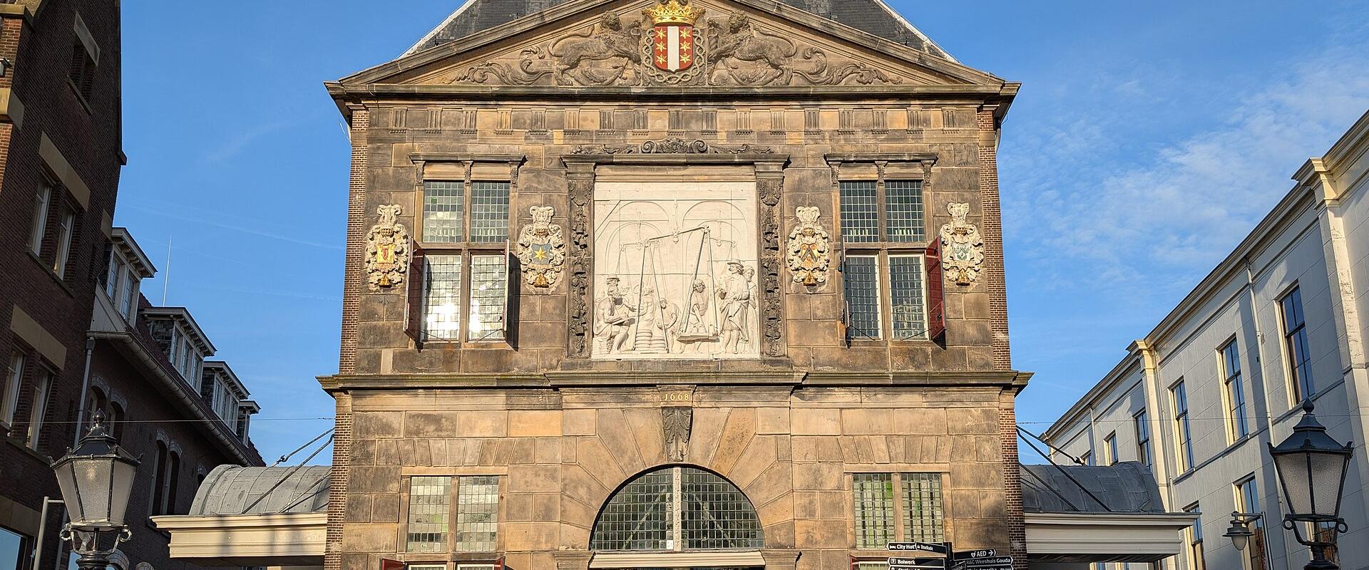 A tall, historic stone weigh house building with a pediment, a large central relief sculpture, and a coat of arms, situated on a brick-paved town square with people walking around.