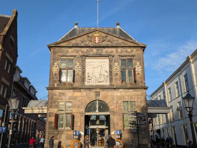 A tall, historic stone weigh house building with a pediment, a large central relief sculpture, and a coat of arms, situated on a brick-paved town square with people walking around.