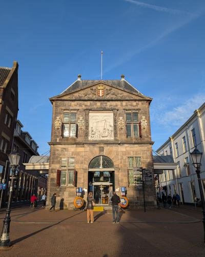 A tall, historic stone weigh house building with a pediment, a large central relief sculpture, and a coat of arms, situated on a brick-paved town square with people walking around.