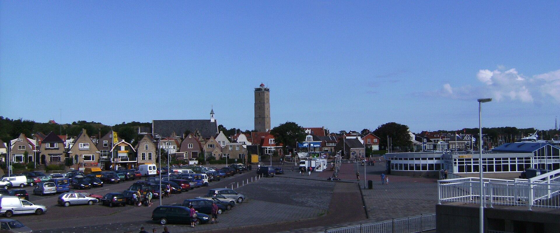 A view of a town square with a parking lot, traditional Dutch houses, a church, and a prominent tower in the background, with a ferry terminal structure visible on the right.