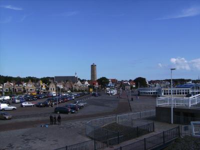 A view of a town square with a parking lot, traditional Dutch houses, a church, and a prominent tower in the background, with a ferry terminal structure visible on the right.