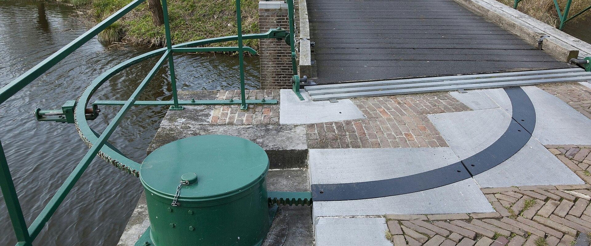 A close-up view of a green-painted drawbridge mechanism, including a winding drum, chain, and railing, situated next to a moat and a wooden bridge deck.