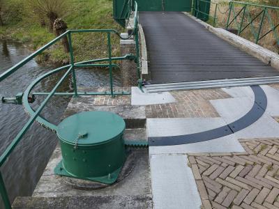 A close-up view of a green-painted drawbridge mechanism, including a winding drum, chain, and railing, situated next to a moat and a wooden bridge deck.