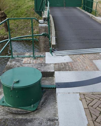 A close-up view of a green-painted drawbridge mechanism, including a winding drum, chain, and railing, situated next to a moat and a wooden bridge deck.
