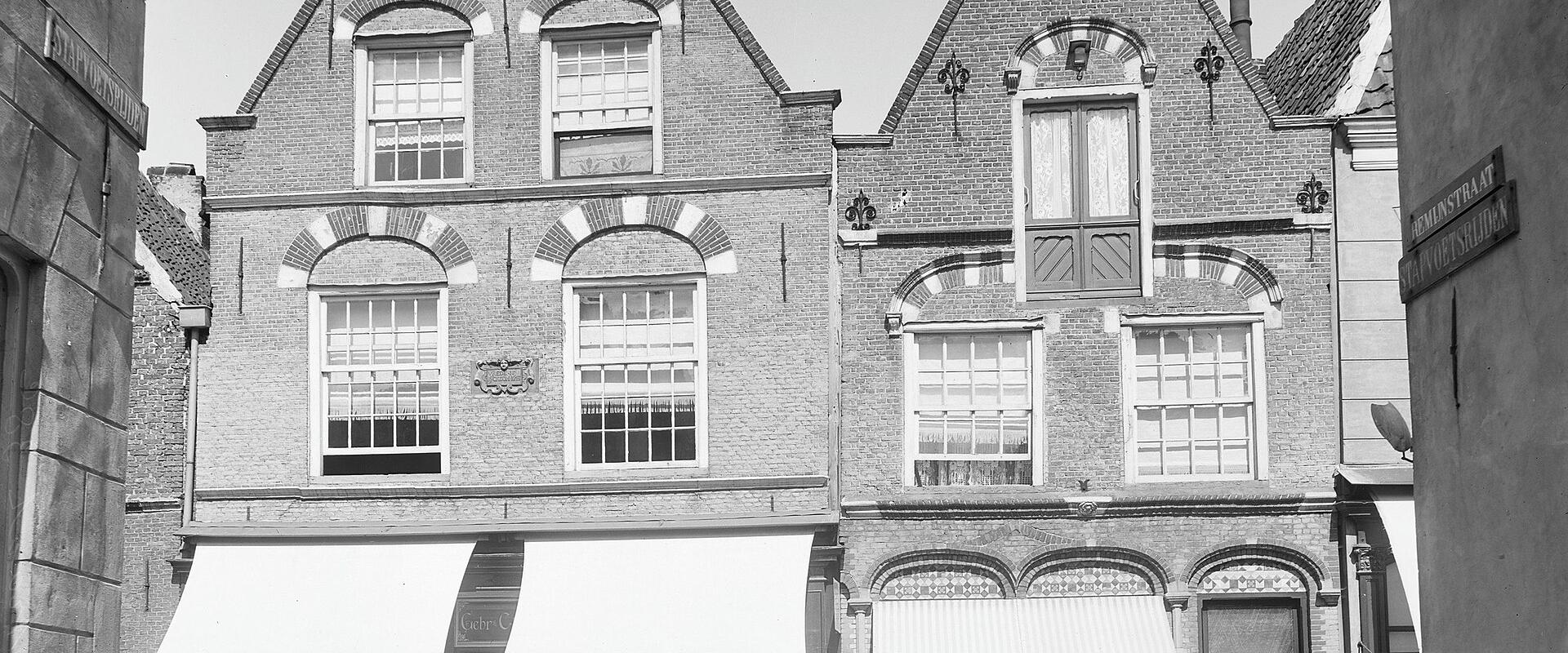 A black and white photograph showing the facades of two adjacent brick buildings with stepped gables and awnings over ground-floor shop windows, with a street sign on the right reading "MUNSTRAAT".