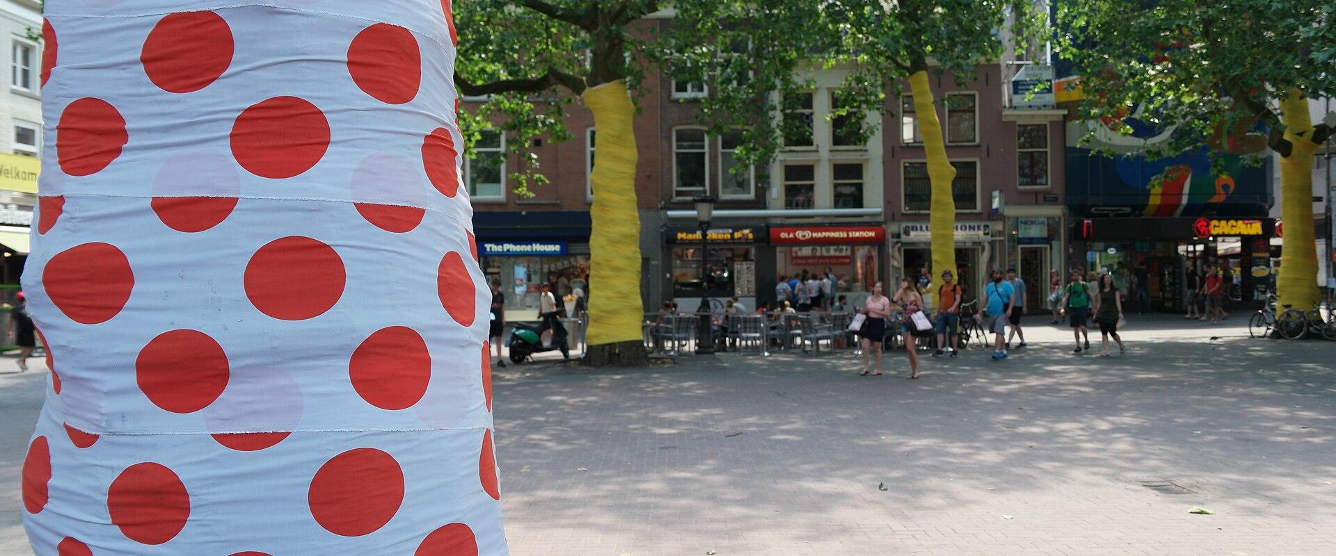 1. A sunlit urban plaza with brick paving, trees wrapped in colorful fabric (red polka dots and yellow), outdoor café seating, pedestrians, and storefronts including "The Phone House" and "GAGAUA" in the background.