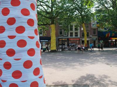 1. A sunlit urban plaza with brick paving, trees wrapped in colorful fabric (red polka dots and yellow), outdoor café seating, pedestrians, and storefronts including "The Phone House" and "GAGAUA" in the background.