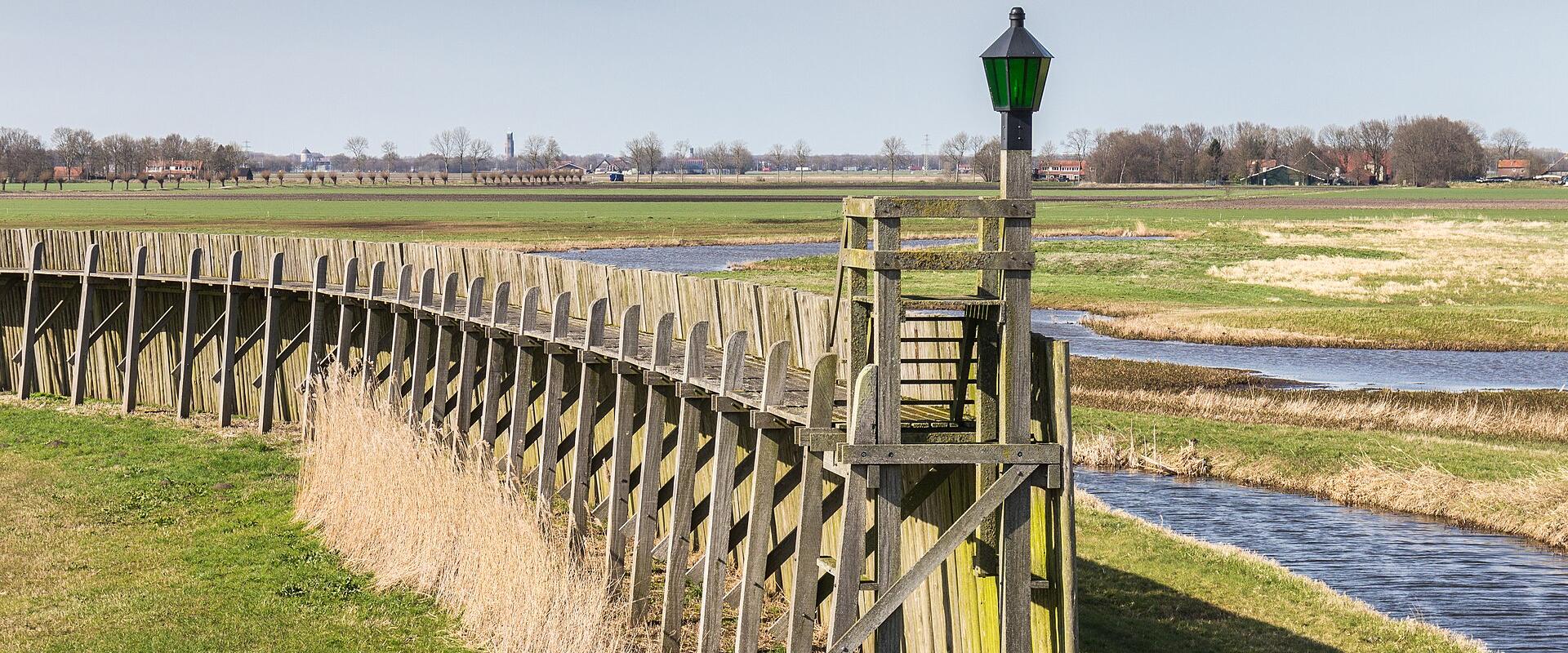 A wooden boardwalk with a green lamp post runs alongside a canal through flat, grassy reclaimed land with distant farmhouses and trees.