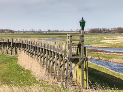 A wooden boardwalk with a green lamp post runs alongside a canal through flat, grassy reclaimed land with distant farmhouses and trees.