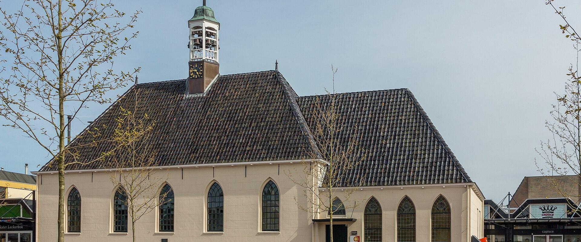 A cream-colored church with a dark tiled roof and a small central bell tower topped with a flag, situated on a paved public square with young trees and adjacent commercial storefronts.