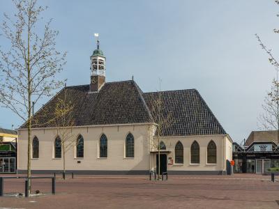 A cream-colored church with a dark tiled roof and a small central bell tower topped with a flag, situated on a paved public square with young trees and adjacent commercial storefronts.