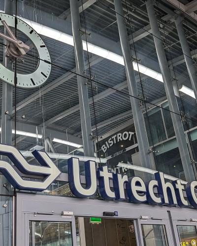 The image shows the interior entrance of a modern train station featuring a large NS (Dutch Railways) logo and a sign reading "Utrecht Centraal" alongside a large hanging clock.