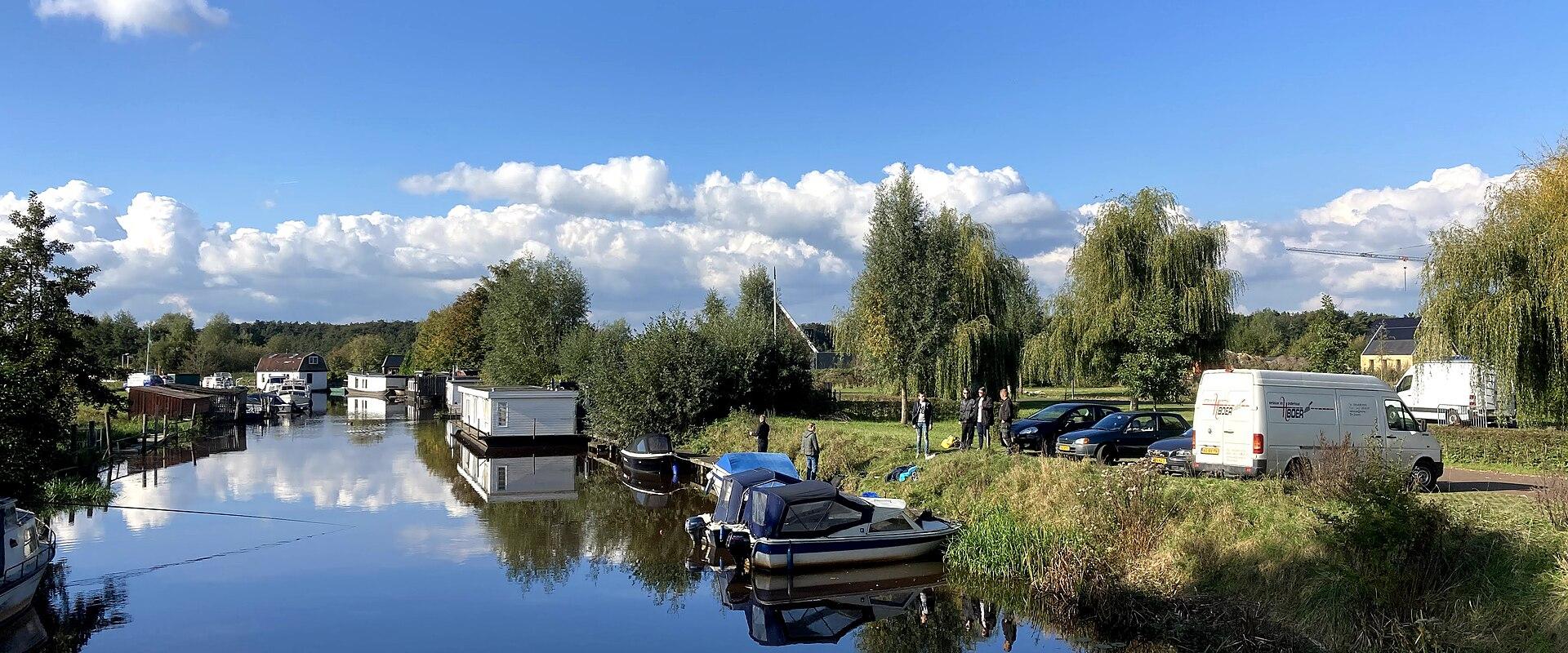 A calm waterway lined with houseboats and small pleasure craft, with a grassy bank on the right where several people and vehicles are parked under trees.