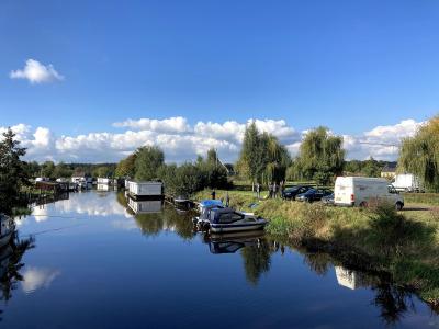 A calm waterway lined with houseboats and small pleasure craft, with a grassy bank on the right where several people and vehicles are parked under trees.