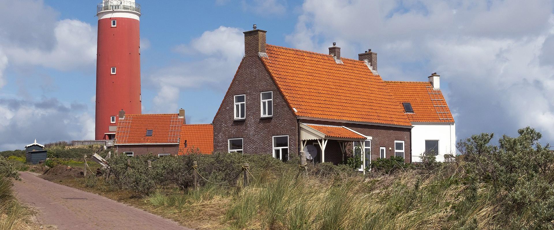 A tall red cylindrical lighthouse with a white lantern room stands behind a traditional brick house with an orange tiled roof, situated on a grassy dune landscape next to a paved path under a cloudy sky.