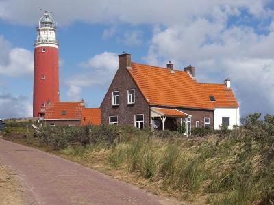 A tall red cylindrical lighthouse with a white lantern room stands behind a traditional brick house with an orange tiled roof, situated on a grassy dune landscape next to a paved path under a cloudy sky.