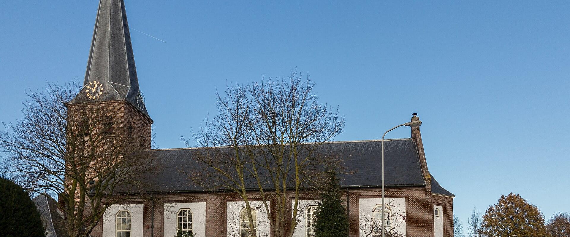 The image shows a brick church with a tall, slender, dark-slated spire, white-painted rectangular panels on the nave, and a street lamp in the foreground.