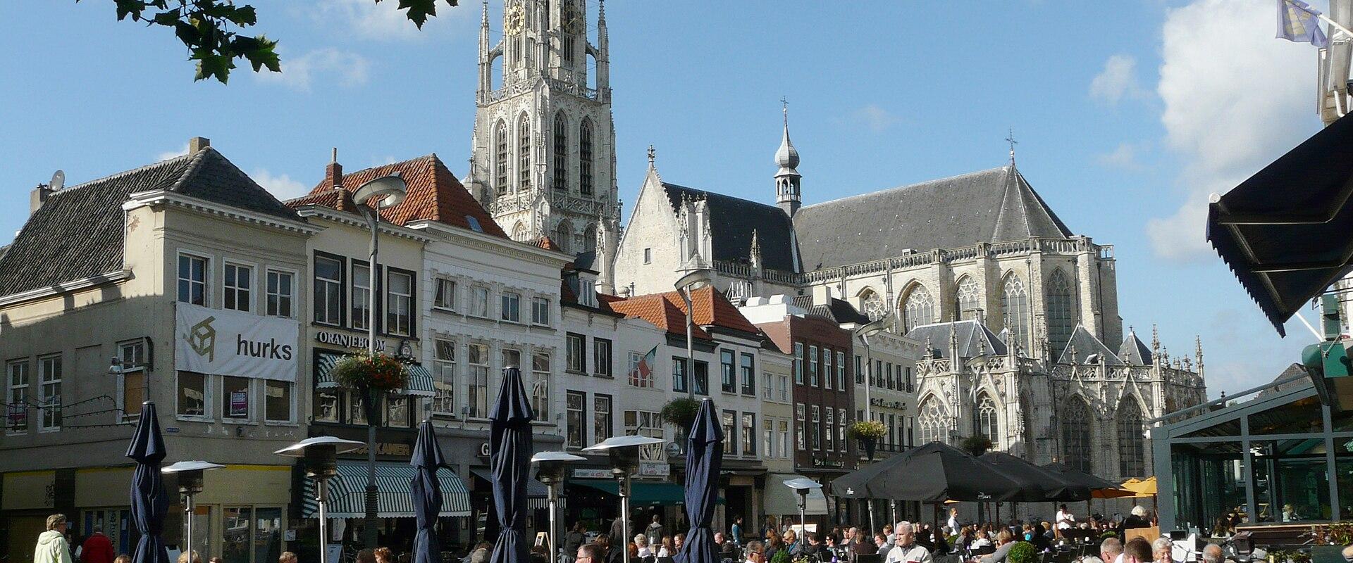 A pedestrian street lined with shops and cafes, featuring outdoor seating, a large tree in the center, and a distinctive church tower visible in the distance at the end of the street.