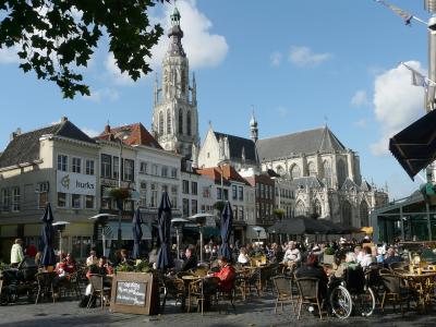 A pedestrian street lined with shops and cafes, featuring outdoor seating, a large tree in the center, and a distinctive church tower visible in the distance at the end of the street.