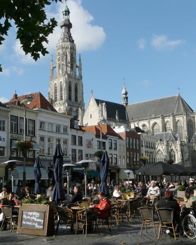 A pedestrian street lined with shops and cafes, featuring outdoor seating, a large tree in the center, and a distinctive church tower visible in the distance at the end of the street.