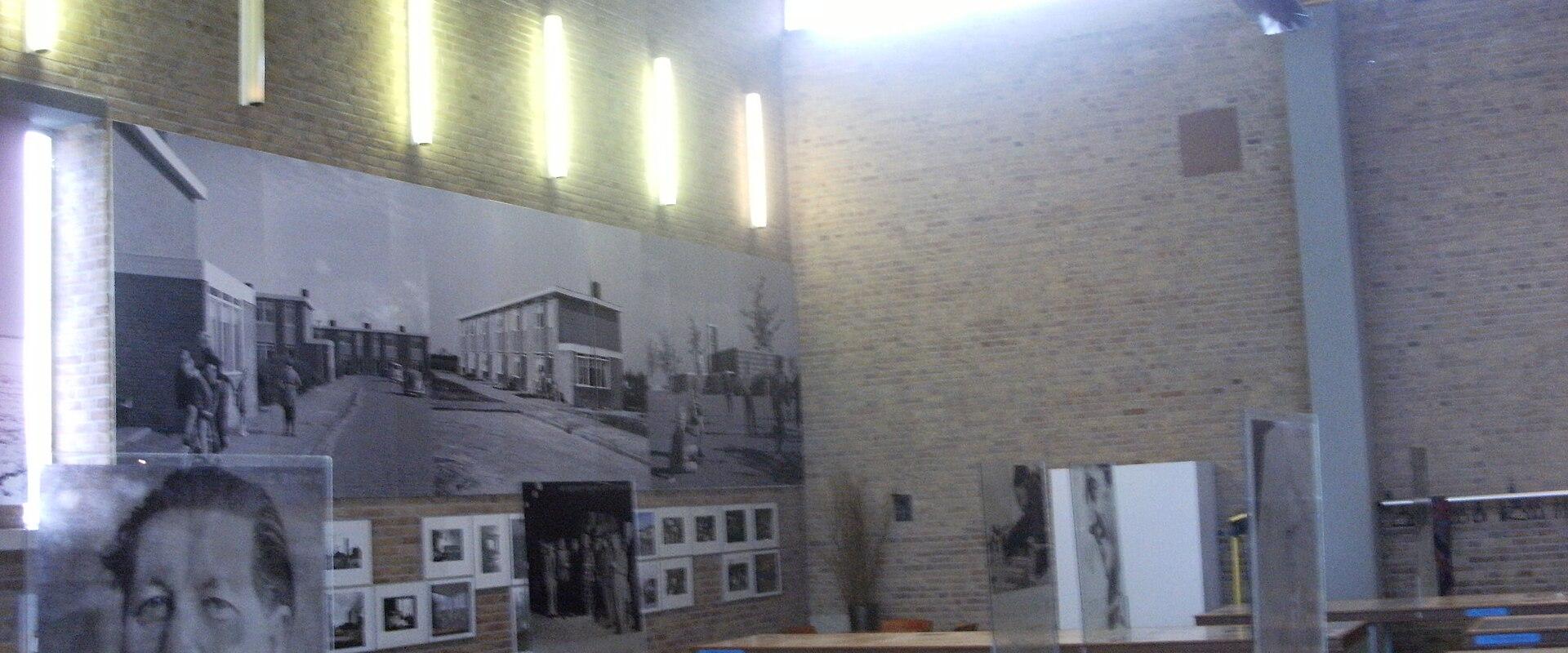 The interior of a museum exhibition space featuring brick walls, vertical light fixtures, display tables, and large black-and-white historical photographs mounted on the walls.