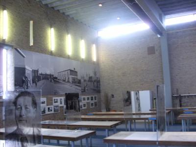 The interior of a museum exhibition space featuring brick walls, vertical light fixtures, display tables, and large black-and-white historical photographs mounted on the walls.